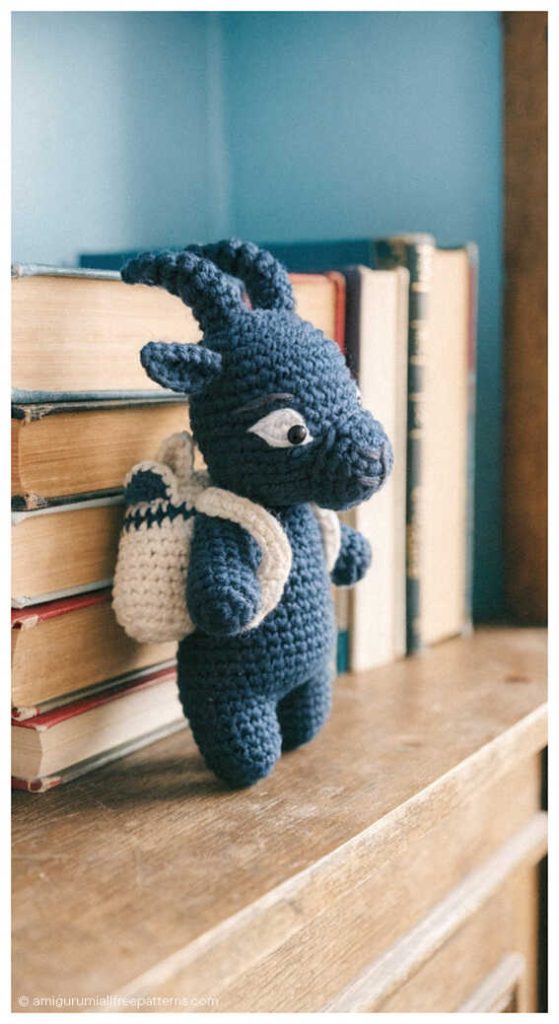 Blue goat amigurumi standing beside stacked books on a wooden shelf, styled as a handmade crochet decor piece for modern nursery or kids room display.