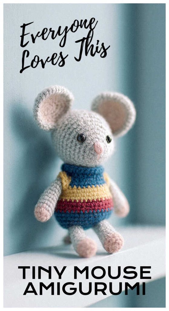 Tiny mouse amigurumi wearing colorful sweater standing against wall, styled as a cute handmade crochet plush toy for nursery decor or gift idea.