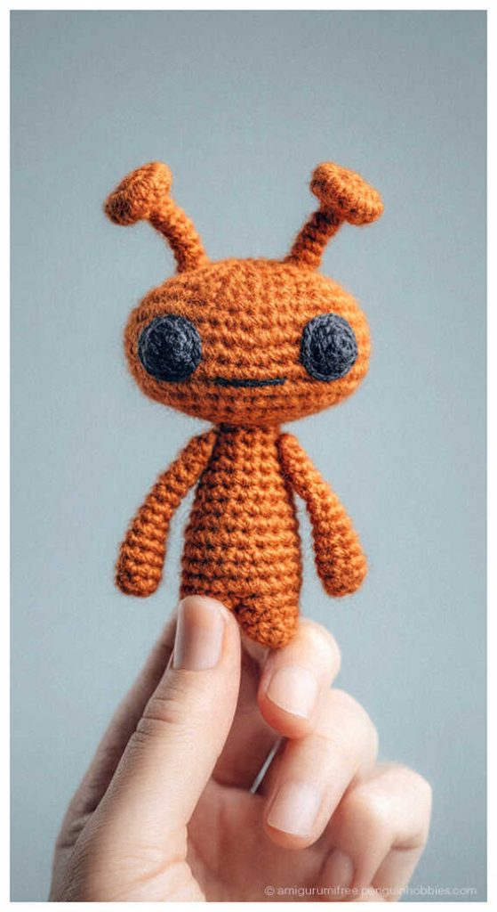 Orange alien amigurumi crochet doll held in hand, styled as a cute handmade space themed plush toy for desk decor or collectible gift.