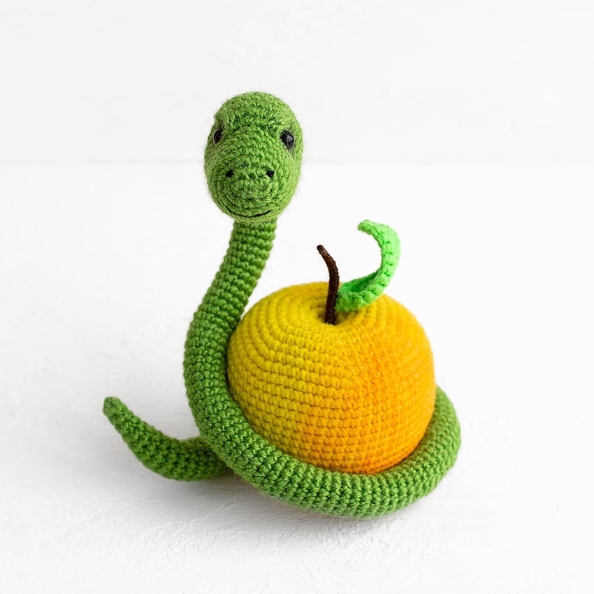 Adorable green crochet snake looking forward while coiled around a handmade yellow amigurumi apple, creative yarn craft idea