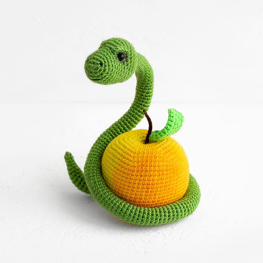 Cute green amigurumi snake wrapped around a yellow crochet apple on a white background, creative handmade plush toy design